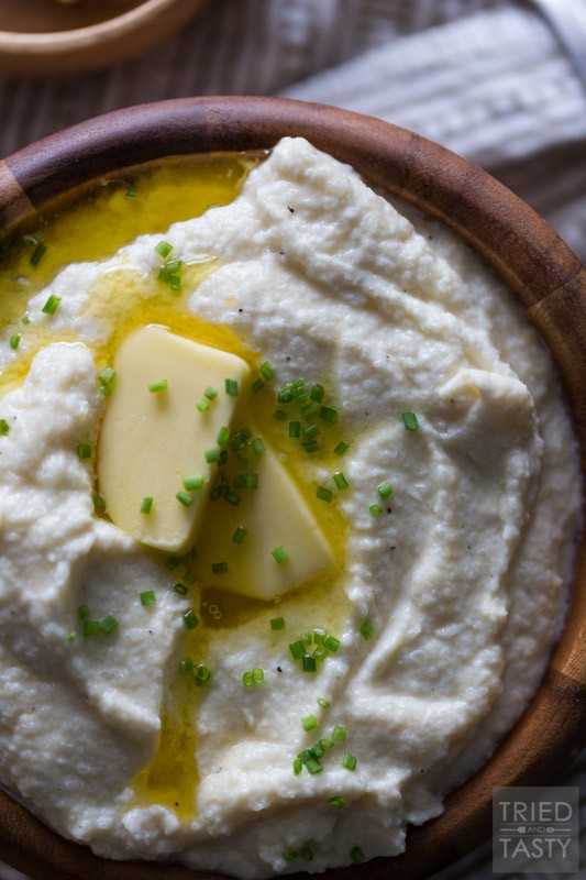 Serving of cauliflower mash in a wooden bowl topped with two pats of butter & chopped chives