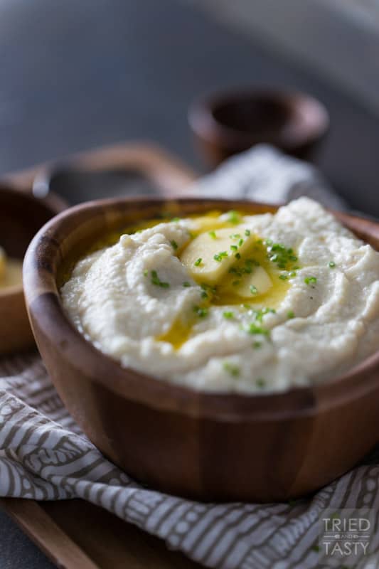 Serving of cauliflower mash in a wooden bowl topped with two pats of butter & chopped chives