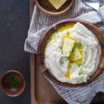 Serving of cauliflower mash in a wooden bowl topped with two pats of butter & chopped chives