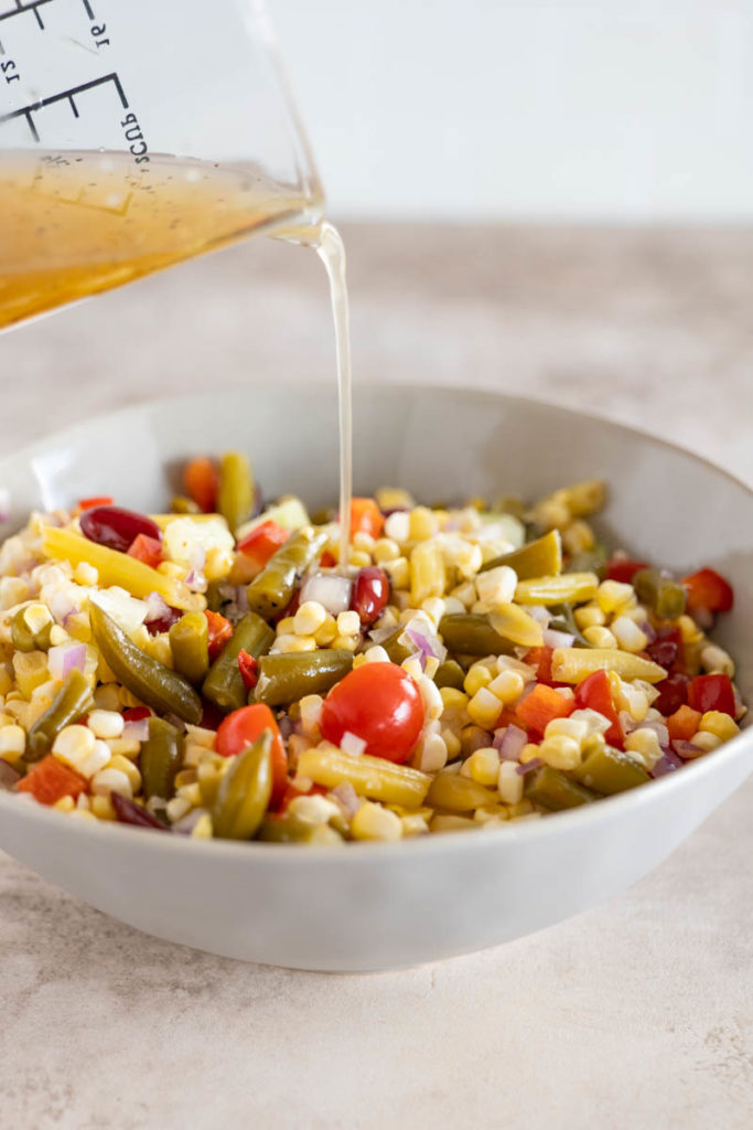 A glass measuring cup drizzling a stream of dressing over a bowl of bean salad.